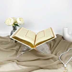 May include: A clear acrylic book stand with a white book resting on top. The book is open and the pages are visible. The stand is on a brown fabric surface.