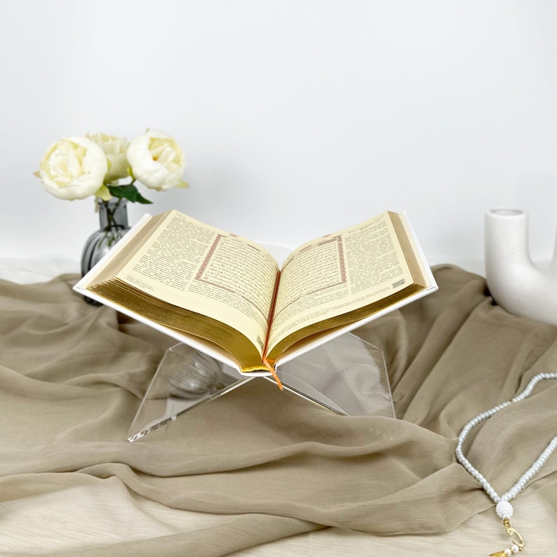 May include: A clear acrylic book stand with a white book resting on top. The book is open and the pages are visible. The stand is on a brown fabric surface.