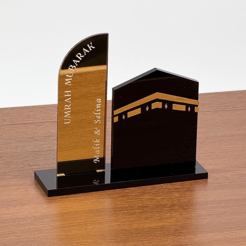 Op de afbeelding: Een zwart en goudkleurig acryl display met de tekst "UMRAH MUBARAK" en de namen "Malik & Selina". De display bevat een goudkleurig, gebogen paneel en een zwarte, geometrische structuur met gouden accenten, alles op een zwarte basis.