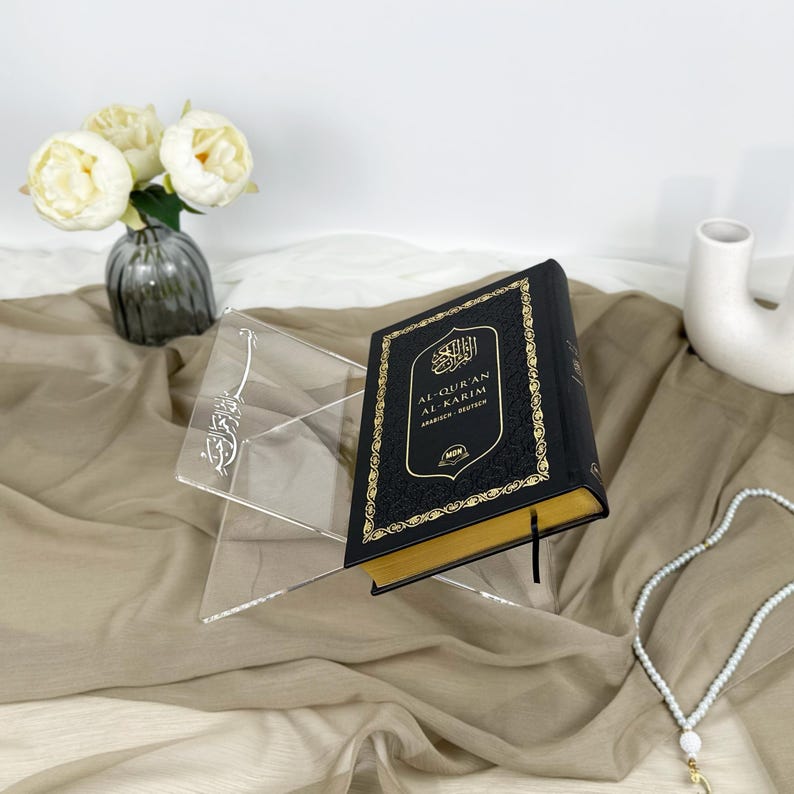 May include: A black Quran book with gold lettering and a gold border on a clear acrylic stand. The book is titled "Al-Quran Al-Karim" in Arabic and English. The stand is on a light brown fabric surface.
