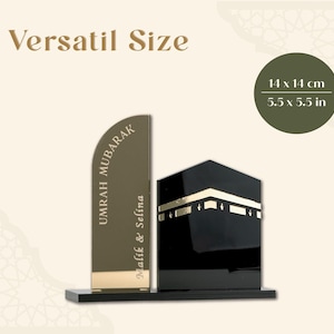 Op de afbeelding: Een decoratieve zwarte en gouden Umrah Mubarak display. De display heeft een gestileerde Kaaba en een verticale plaat met de tekst "UMRAH MUBARAK" en "Malik & Selina". Afmetingen: 14 x 14 cm.