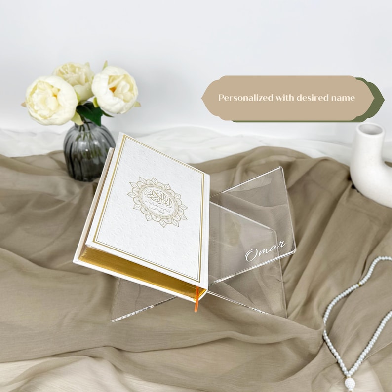 May include: A white book with gold accents and Arabic calligraphy on the cover is displayed on a clear acrylic stand. The stand is personalized with the name "Omar".