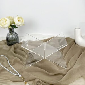 May include: A clear acrylic book stand with a personalized inscription that reads "Omar". The stand is designed to hold books or other items upright.