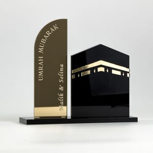 Op de afbeelding: Een decoratief islamitisch kunstwerk. Het toont een goudkleurige, gebogen plaat met de woorden "UMRAH MUBARAK" en "Malik & Selina" gegraveerd. Daarnaast een zwarte, kubusvormige structuur met gouden accenten, alles op een zwarte basis.