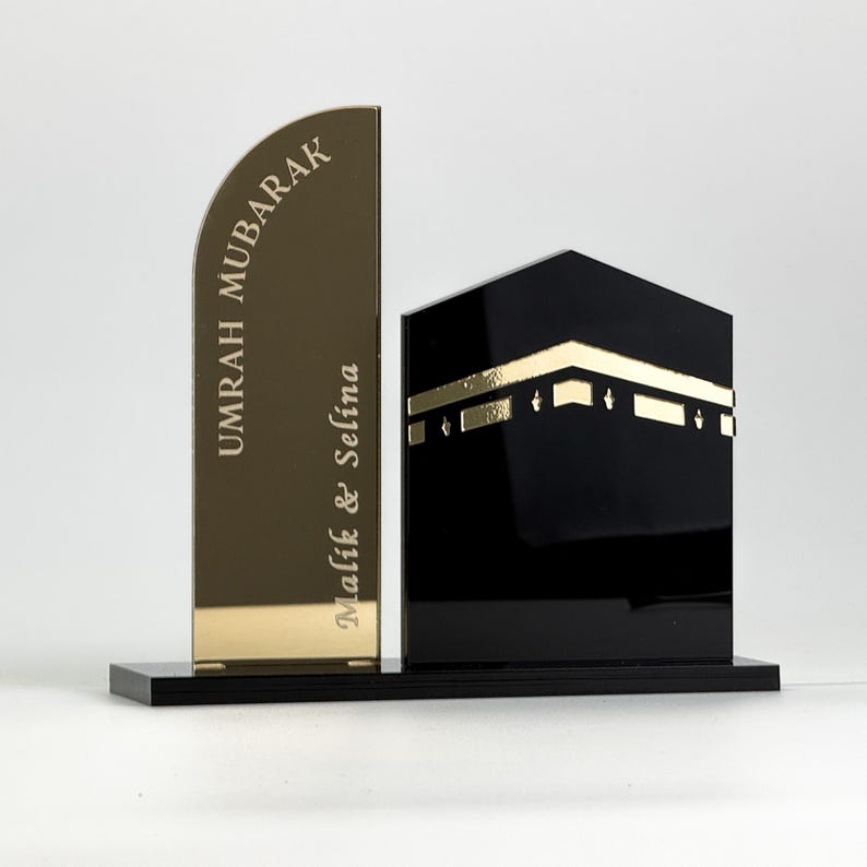 Op de afbeelding: Een decoratief islamitisch kunstwerk. Het toont een goudkleurige, gebogen plaat met de woorden "UMRAH MUBARAK" en "Malik & Selina" gegraveerd. Daarnaast een zwarte, kubusvormige structuur met gouden accenten, alles op een zwarte basis.
