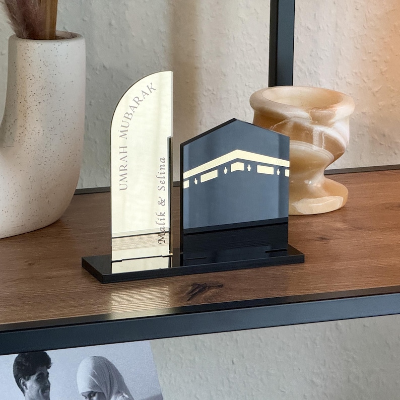 Op de afbeelding: Een decoratieve zwarte en gouden acryl display met een gestileerde weergave van de Kaaba en een verticale plaat met de tekst "UMRAH MUBARAK" en de namen "Malik & Selina". De display staat op een zwarte basis.