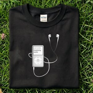 May include: Black t-shirt with a white graphic of a digital music player and earbuds. The player's screen reads "NOW PLAYING mommy asmr". The earbuds' white cords are draped across the shirt.