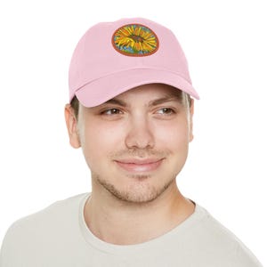 May include: A pink baseball cap with a sunflower patch on the front. The sunflower is yellow with a brown centre and green leaves. The patch is surrounded by a brown circle.