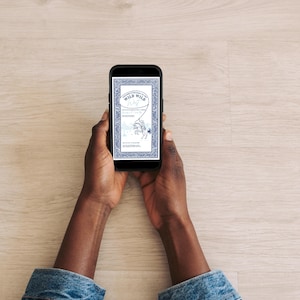 Close-up of hands holding a smartphone displaying the blue “Wild Wild West” invitation template. The Western invitation is clearly visible on the phone screen against a light wood background, showing mobile-friendly format.