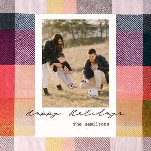 May include: A family of three, a woman, a man, and a young child, are playing with a soccer ball in a field. The photo is framed in a white border with the text "Happy Holidays" and "The Hamiltons" below the photo. The background is a colorful plaid pattern.