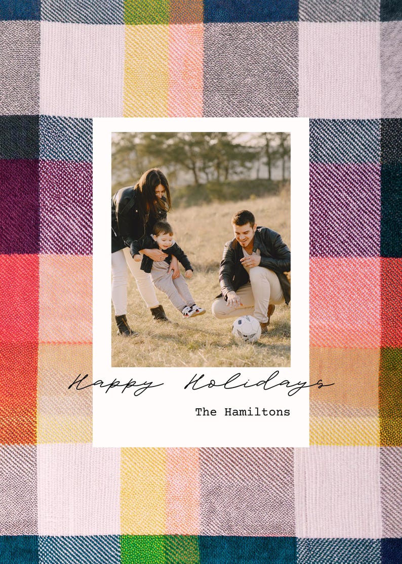 May include: A family of three, a woman, a man, and a young child, are playing with a soccer ball in a field. The photo is framed in a white border with the text "Happy Holidays" and "The Hamiltons" below the photo. The background is a colorful plaid pattern.