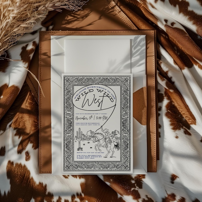 Western-themed invitation mockup with a black and cream “Wild Wild West” design placed inside a tan leather tray on a cowhide-print fabric background. The invitation features a detailed paisley border and cowboy illustration with desert scenery, creating a rustic, elegant Western aesthetic ideal for rodeo or cowboy-themed events.