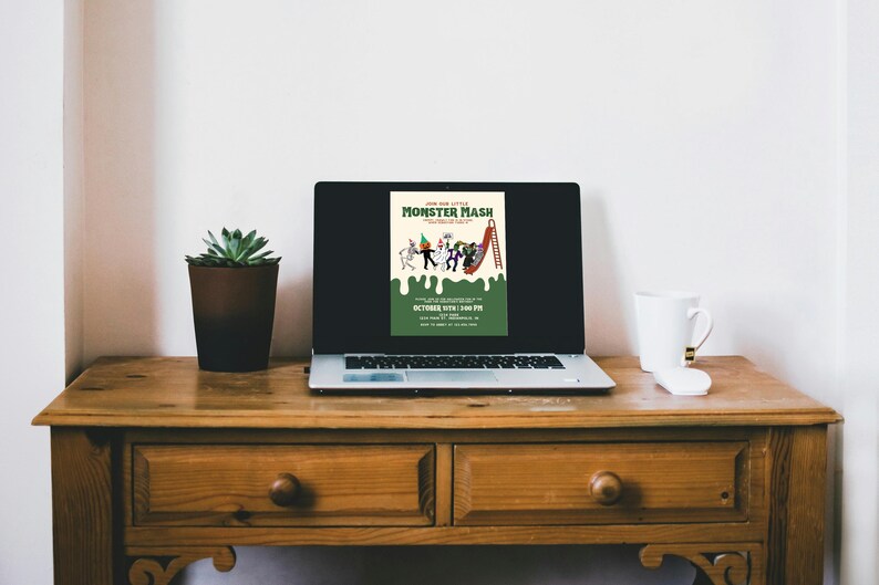 May include: A laptop displays a Halloween-themed poster with the text "Join Our Little Monster Mash." The poster features cartoon monsters and a slide. A potted succulent and a white mug with a black stripe sit on a wooden desk.