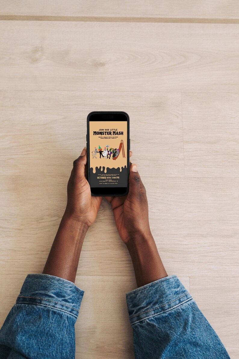 Hands holding a smartphone that displays a digital Monster Mash party invitation on a light wooden table background.