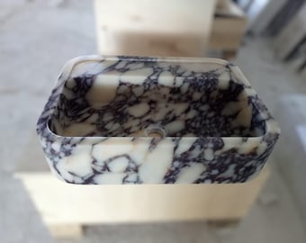Puede incluir: Un lavabo de mármol blanco con vetas moradas. El lavabo tiene una forma rectangular con bordes redondeados.