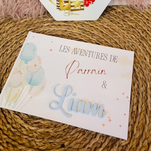 Peut inclure: Une carte hexagonale blanche avec des lettres dorées et des accents floraux rouges, indiquant "Lyma ginative". En dessous, deux sacs à provisions dorés sont représentés. Un livre avec le texte "LES AVENTURES DE Parrain & Liam" est également visible.