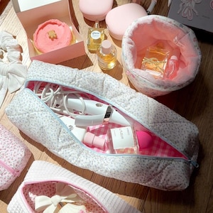 May include: A white quilted makeup bag with a floral print and pink trim. The bag is open and filled with various beauty products, including a curling iron, a bottle of perfume, and a makeup sponge.