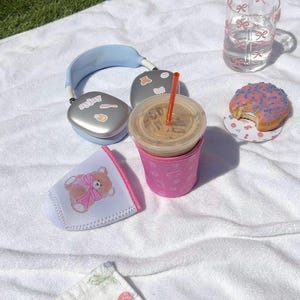 May include: A picnic scene with a pink iced donut, a glass of water, a pink insulated cup holder with a teddy bear design, a silver pair of headphones, and a cup of iced coffee with a straw.
