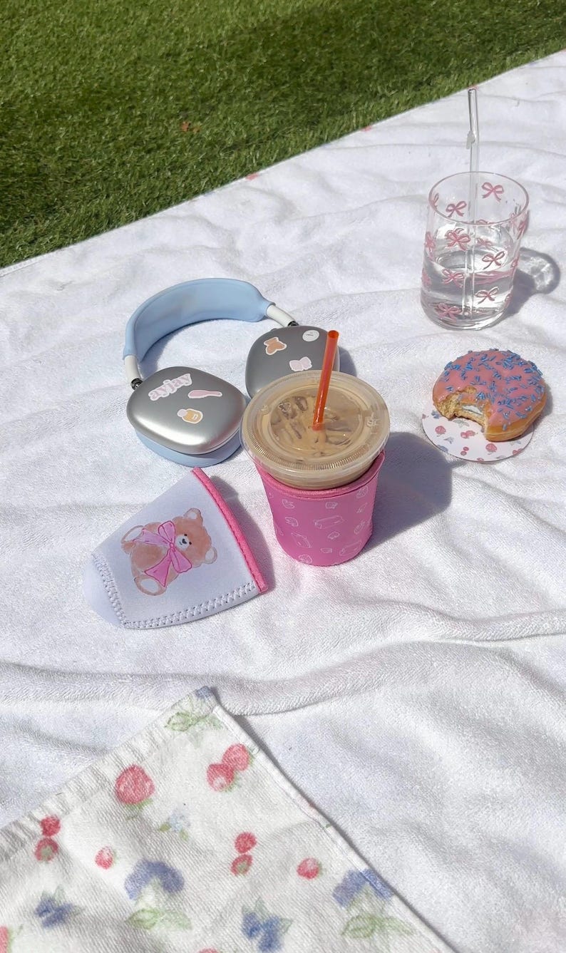 May include: A picnic scene with a pink iced donut, a glass of water, a pink insulated cup holder with a teddy bear design, a silver pair of headphones, and a cup of iced coffee with a straw.