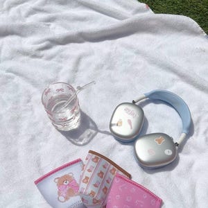 May include: Three pink and white patterned drink koozies with a bear design, a clear glass with a pink bow pattern and a blue and silver wireless headset with the word "anjaya" on it.