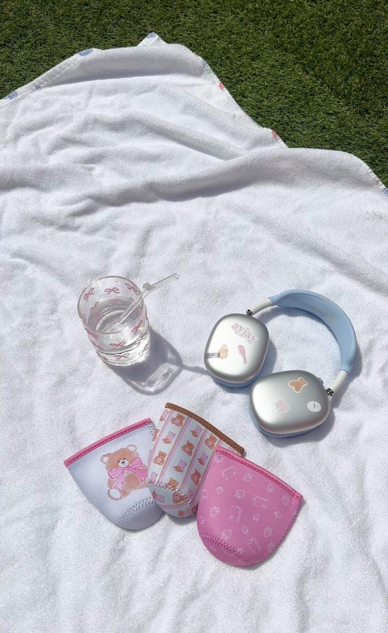 May include: Three pink and white patterned drink koozies with a bear design, a clear glass with a pink bow pattern and a blue and silver wireless headset with the word "anjaya" on it.