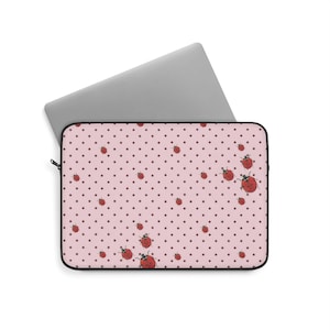 Może przedstawiać: Etui na laptopa z jasnoróżowym tłem, wzorem w małe ciemnobrązowe kropki oraz rozproszonymi ilustracjami czerwonych biedronek. Etui posiada czarne wykończenie z zamkiem błyskawicznym i częściowo przykrywa srebrny komputer przenośny.