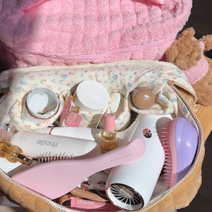 Può includere: Una borsa in tessuto trapuntato rosa contenente una pochette a fiori piena di prodotti di bellezza, tra cui un asciugacapelli bianco, una spazzola rosa chiaro, una spazzola districante viola, un piccolo flacone d'olio e due tubetti marchiati rhode.