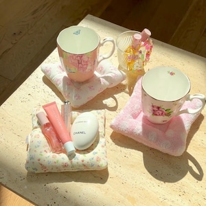 May include: Two floral teacups with pink and white floral patterns, one on a pink and white floral coaster and the other on a pink and white towel coaster.  A small white jar with the word "CHANEL" on it, a pink and white floral patterned coaster, and two pink lip glosses are on a white and yellow floral patterned coaster.