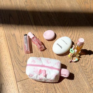 Op de afbeelding: Een witte gewatteerde toilettas met roze strikken, een roze lipgloss-buisje, een roze en doorzichtig flesje parfum, een witte ronde container met een roze deksel, een witte tube handcrème met de tekst "LA CREME MAIN CHANEL" en een klein doorzichtig flesje parfum met een roze en groene kralenketting.