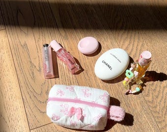 Puede incluir: Una bolsa de maquillaje acolchada blanca con lazos rosas, un tubo de brillo labial rosa, una botella de perfume rosa y transparente, un recipiente redondo blanco con una tapa rosa, un tubo de crema de manos blanco con el texto "LA CREME MAIN CHANEL" y una pequeña botella de perfume transparente con una cadena de cuentas rosas y verdes.