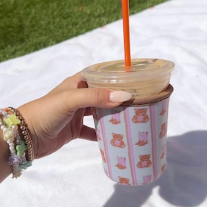 May include: A pink and white insulated cup sleeve with a repeating pattern of teddy bears. The sleeve is on a white towel and has an orange straw sticking out of the top.