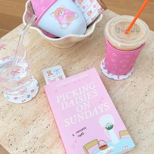 May include: A pink book titled "Picking Daisies on Sundays: A Romance Novel" with a floral illustration on the cover. The book is on a light brown table with a pink and white patterned coaster, a glass of water with a pink straw, a pink and white patterned cup holder with a cup of coffee, and a pink and white patterned cup holder with a teddy bear illustration.