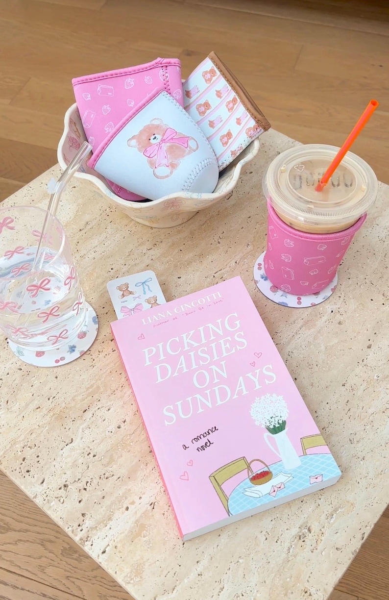 May include: A pink book titled "Picking Daisies on Sundays: A Romance Novel" with a floral illustration on the cover. The book is on a light brown table with a pink and white patterned coaster, a glass of water with a pink straw, a pink and white patterned cup holder with a cup of coffee, and a pink and white patterned cup holder with a teddy bear illustration.