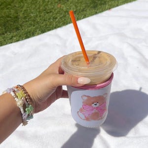 May include: A white insulated cup with a pink trim and a cartoon teddy bear with a pink bow. The cup is holding a cold beverage with a straw.