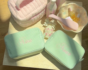 May include: A collection of beauty and personal care items, including a pink quilted cosmetic bag, a floral-patterned tumbler, and two green striped cosmetic bags embroidered with the name "Nina". Other items include a perfume bottle, a brush, and small bottles.