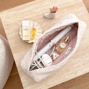 May include: A white, fluffy cosmetic bag with a floral interior holds a curling iron, skincare products, and a small Chanel cream container. A small ceramic boot holds pink matches. Other beauty products are displayed on a decorative plate.