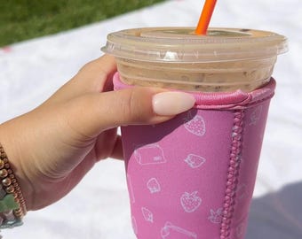 May include: A pink insulated cup sleeve with a white strawberry pattern. The sleeve is on a white surface and has a straw sticking out of the top.