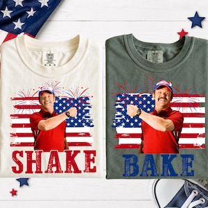 May include: Two t-shirts, one cream and one dark gray, each featuring a graphic of a man in a red polo shirt and cap against an American flag and fireworks background. The cream shirt has the word SHAKE in red, and the gray shirt has BAKE in blue.
