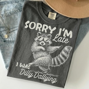 May include: A charcoal gray t-shirt featuring a white graphic of a raccoon wearing sunglasses. The text reads SORRY I'M Late and I WAS Dilly Dallying. The shirt has a crew neckline and a Comfort Colors brand label printed on the inside collar area.