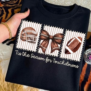 May include: A black sweatshirt featuring three rectangular patches with a brown and white striped background. The patches display a brown football helmet, a brown bow, and a brown football. The text 'Tis the Season for Touchdowns' is printed below.