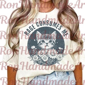 May include: Cream-colored t-shirt featuring a distressed graphic of a kitten surrounded by flowers. The text "RAGE CONSUMES ME" is printed in a circular design above the kitten. The shirt is made of a soft material.