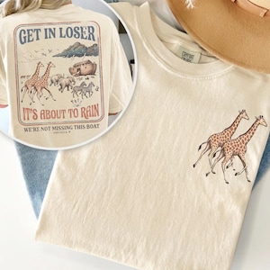 Peut inclure: Un t-shirt couleur crème avec un motif de deux girafes sur le devant. Le dos présente une grande illustration de girafes, d'éléphants et de zèbres s'approchant d'une arche avec le texte : GET IN LOSER IT'S ABOUT TO RAIN WE'RE NOT MISSING THIS BOAT GENESIS 6-9.