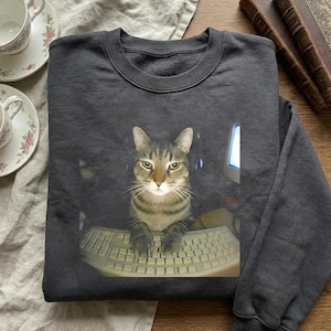 Puede incluir: Sudadera gris oscuro con un gato atigrado con auriculares y sentado frente a un teclado de ordenador. Un monitor de ordenador es visible en el fondo. La sudadera está doblada. El gato es el foco principal.