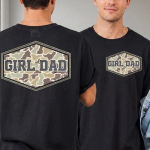 May include: A black t-shirt featuring a large hexagonal graphic on the back and a smaller matching version on the front left chest. The graphic contains a camouflage pattern in shades of green, tan, and brown with the text GIRL DAD in bold.