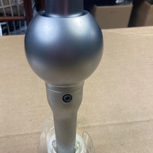 May include: A silver gear shift knob with a round top and a black screw. The knob is mounted on a clear plastic base.