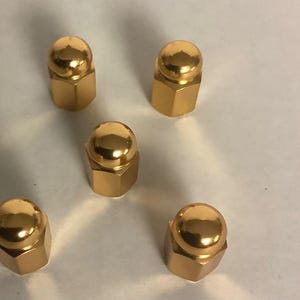 May include: Five gold-colored acorn nuts are arranged on a light surface. Each nut has a rounded top and a hexagonal base. The shiny finish reflects light, creating highlights on the surface of each nut.
