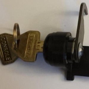 May include: A black and gold console lock with two keys and a latch. The keys are attached to a ring and have the word "CONSOLE" printed on them. The lock is black and has a silver screw.