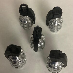 May include: Five silver-colored grenade-shaped objects with black tops are arranged on a white surface. The grenades have a ribbed body and a black lever and pin. The objects are evenly spaced and appear to be made of metal.