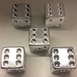 May include: Five silver dice with deeply recessed dots are arranged on a light gray surface. The dice are evenly spaced, with each die showing a different face. The dice are made of a shiny, metallic material.
