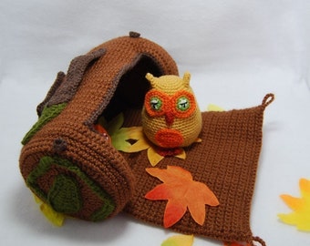 May include: A crocheted owl toy sits inside a crocheted log cabin with a brown crocheted mat and fall-colored leaves scattered around.